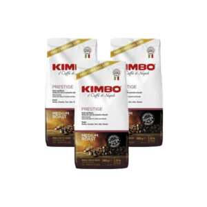 Κόκκοι καφέ Prestige KIMBO, 3 συσκευασίες του 1 kg, λευκή και κόκκινη συσκευασία, λογότυπο στο επάνω μέρος, ορατοί κόκκοι καφέ.